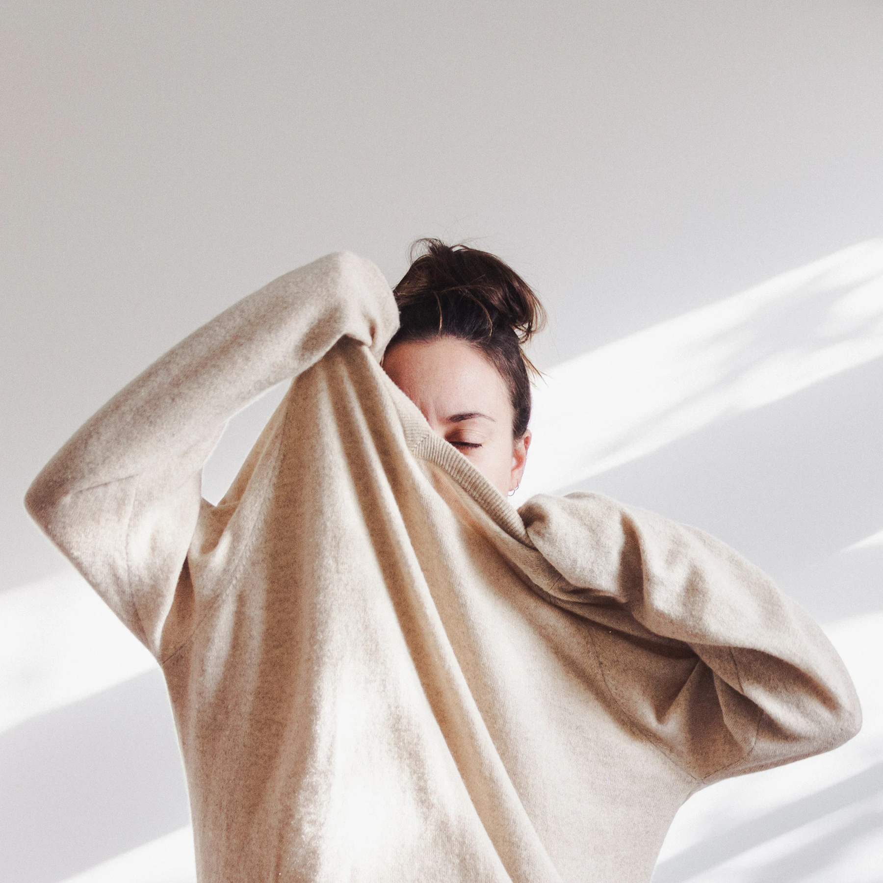 Woman pulling an oat knit sweater over her face in soft light