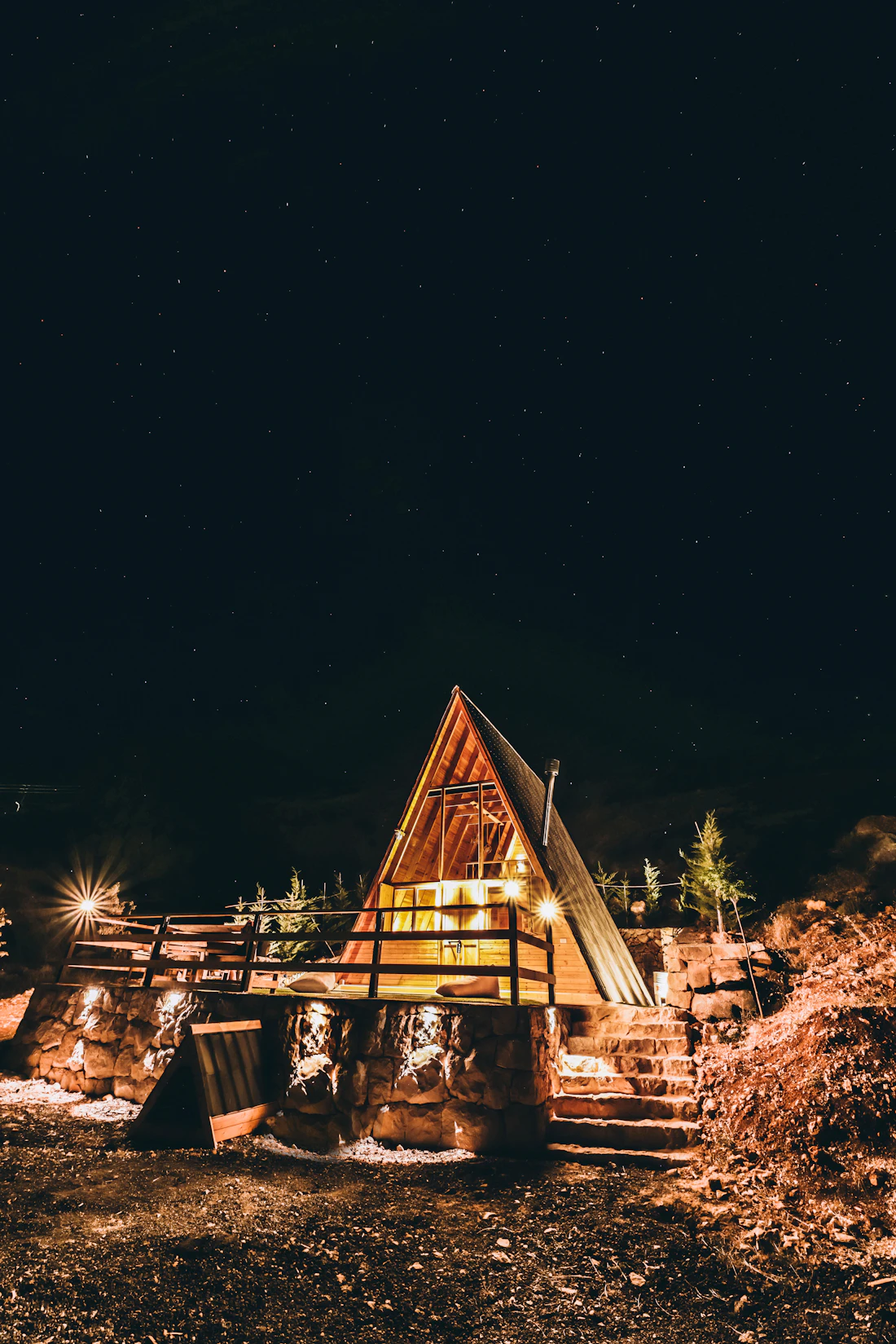 A small A-frame cabin lit from inside, with a deck, at night