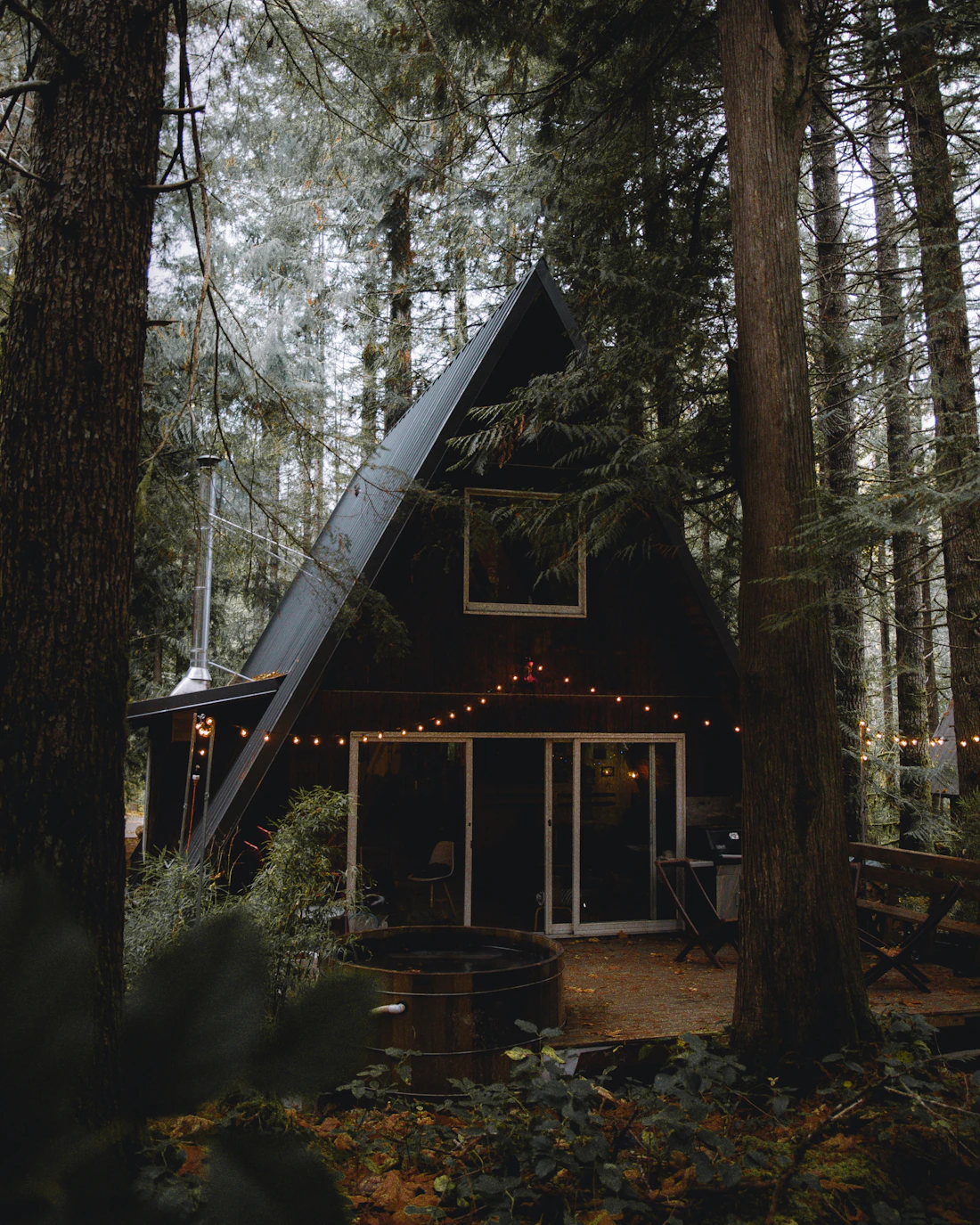 A dark A-frame cabin among tall trees, with warm lights on