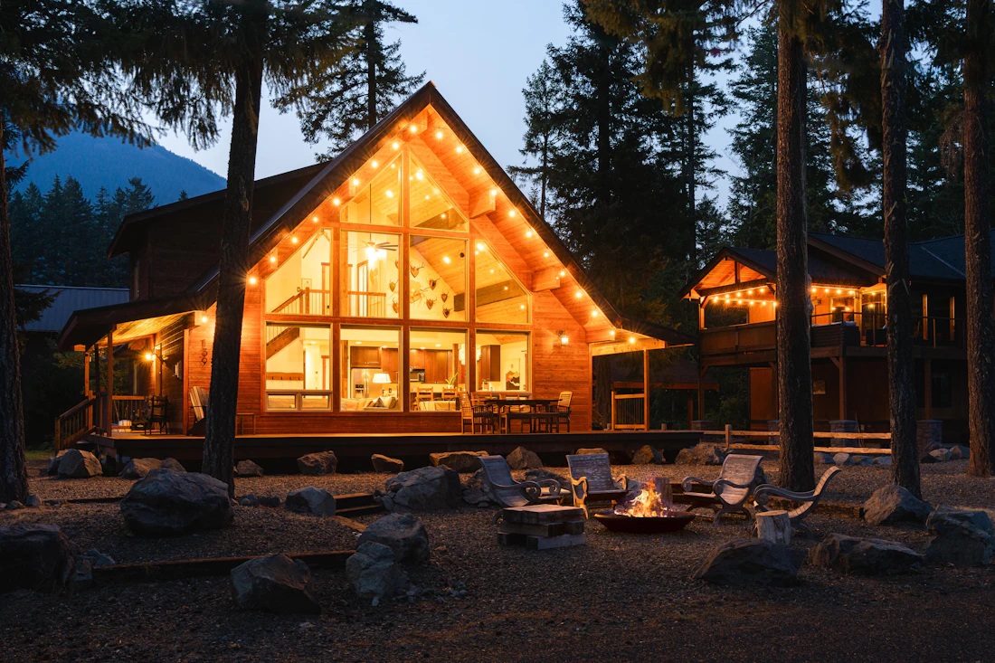 A large cabin with a glass front glowing at dusk, a fire pit in front