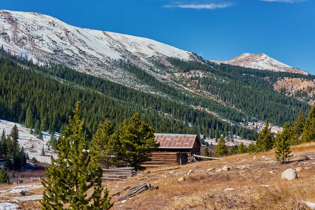 An old log cabin in a mountain valley, snow on the peaks