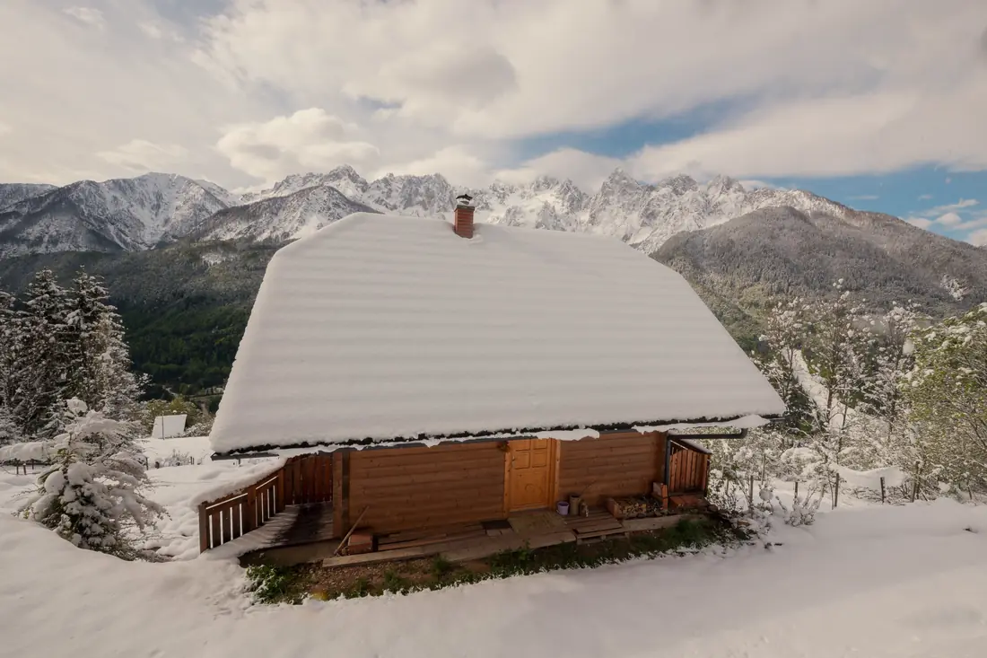 A small wooden cabin under deep snow, with mountains behind