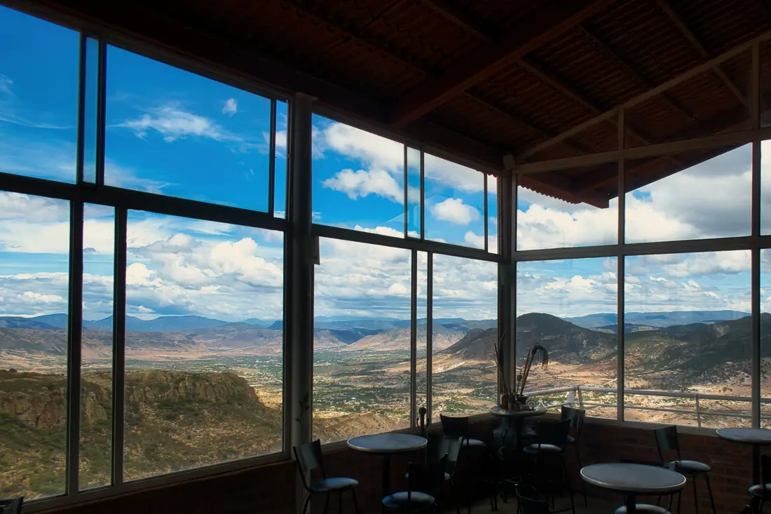 A glass walled terrace with small tables, looking over a wide valley